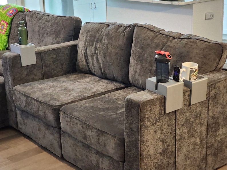May include: Three light grey rectangular sofa armrest trays designed to drape over the sides of a sofa. Each tray features a recessed circular cutout for holding beverages or items like a black shaker bottle, a white mug, or a remote control.