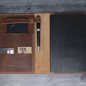 May include: A brown leather notebook cover with a pen holder, a pocket for a phone, and a card slot. The cover is open to reveal a black notebook.