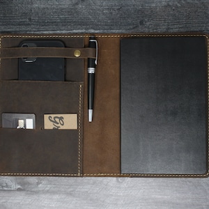May include: A brown leather portfolio with a black notebook, a pen holder, a phone pocket, and a card slot. The portfolio has a brown leather flap with a brass snap closure.