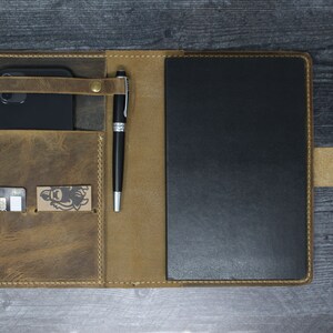 May include: A brown leather notebook cover with a pen holder, a phone pocket, and a card slot. The cover is open to reveal a black notebook.