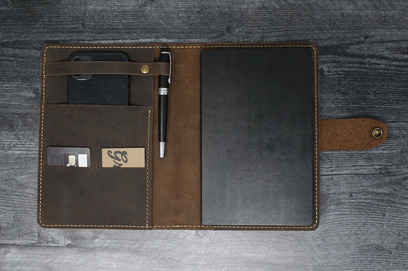 May include: A brown leather portfolio with a black notebook, a pen holder, a phone pocket, and a card slot. The portfolio has a brown leather flap with a brass snap closure.