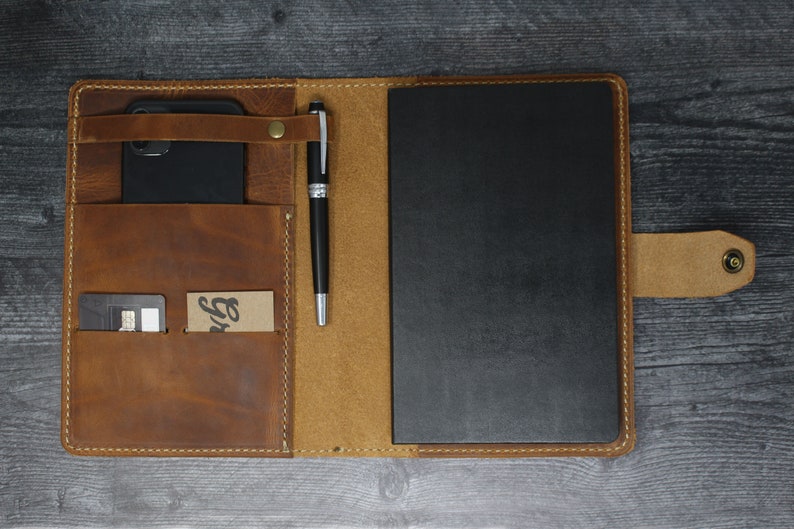 May include: A brown leather portfolio with a black notebook, a pen, a phone, and a card holder. The portfolio has a flap with a snap closure and a strap to hold the pen.