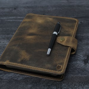 May include: A brown leather journal with a pen on top. The journal has a flap closure and a stitched edge. The pen is black with a silver clip.