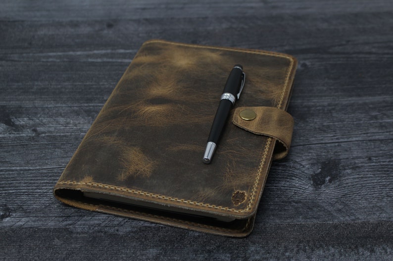 May include: A brown leather journal with a pen on top. The journal has a flap closure and a stitched edge. The pen is black with a silver clip.