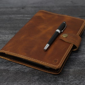 May include: A brown leather journal with a pen tucked inside the flap. The journal has a stitched edge and a small metal clasp.