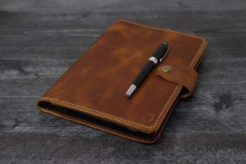 May include: A brown leather journal with a pen tucked inside the flap. The journal has a stitched edge and a small metal clasp.