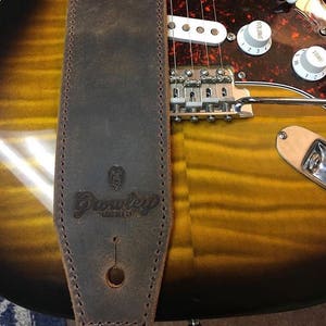 May include: A brown leather guitar strap with the word "Grooveley" embossed on it. The strap is attached to a guitar with a gold finish.
