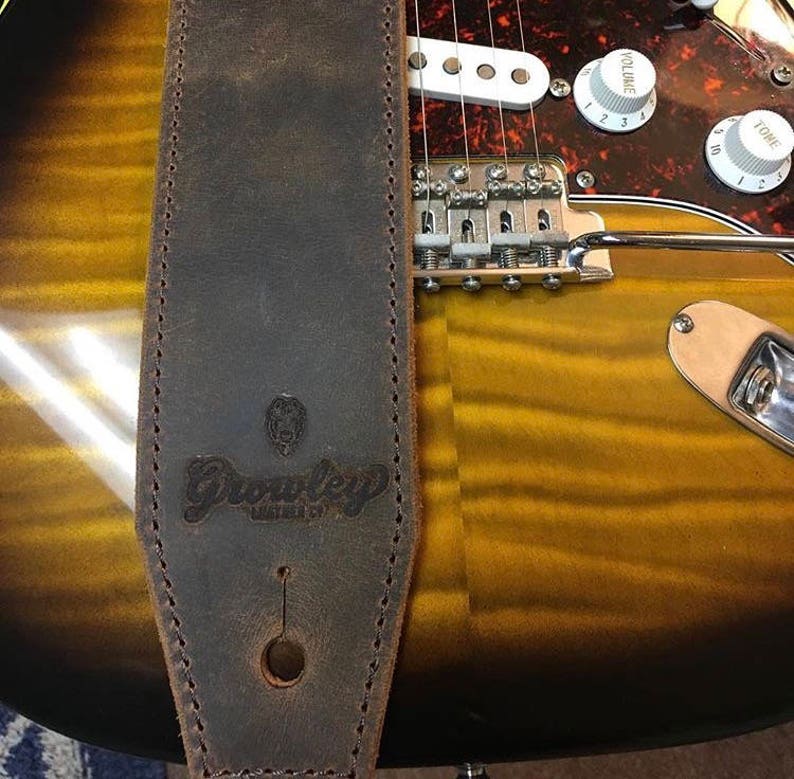 May include: A brown leather guitar strap with the word "Grooveley" embossed on it. The strap is attached to a guitar with a gold finish.