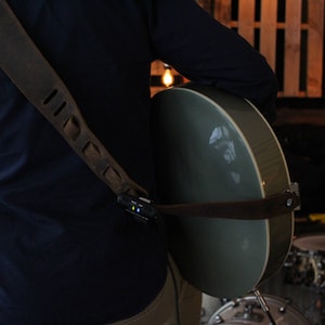 May include: A person wearing a dark blue shirt with a brown leather guitar strap and a grey acoustic guitar. The guitar is slung over the person's shoulder and the strap has a small black device attached to it.