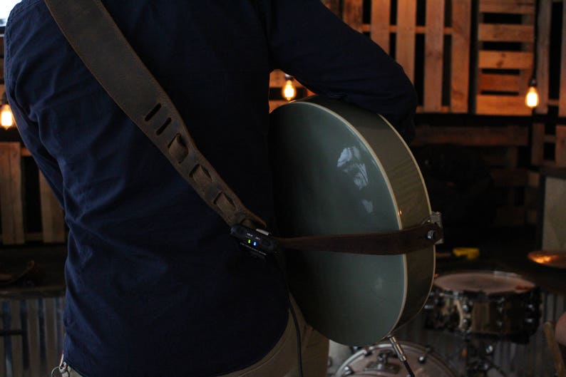 May include: A person wearing a dark blue shirt with a brown leather guitar strap and a grey acoustic guitar. The guitar is slung over the person's shoulder and the strap has a small black device attached to it.