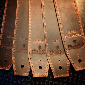 May include: Five brown leather guitar straps with different names embossed on them. The names are LCS, RJD, RCM, NP, and DAVID FRANCISCO. Each strap has a small hole for a strap button and the Growley Leather Co. logo is embossed on each strap.