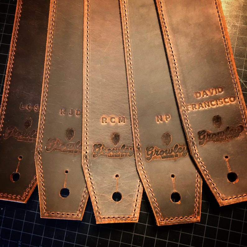 May include: Five brown leather guitar straps with different names embossed on them. The names are LCS, RJD, RCM, NP, and DAVID FRANCISCO. Each strap has a small hole for a strap button and the Growley Leather Co. logo is embossed on each strap.