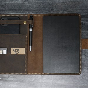 May include: A brown leather portfolio with a black notebook, a pen holder, a phone pocket, and a card slot. The portfolio has a brown leather strap with a gold metal button.