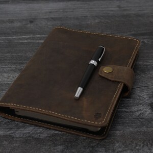 May include: A brown leather-bound journal with a pen on top. The journal has a flap closure with a brass button.