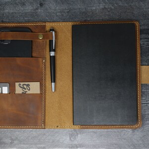 May include: A brown leather portfolio with a black notebook, a pen, a phone, and a card holder. The portfolio has a flap with a snap closure and a strap to hold the pen.