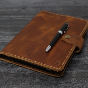 May include: A brown leather journal with a pen tucked inside the flap. The journal has a stitched edge and a small metal clasp.