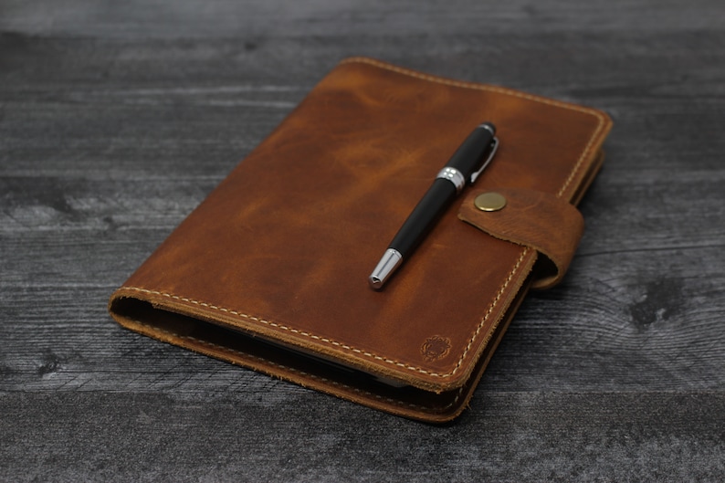 May include: A brown leather journal with a pen tucked inside the flap. The journal has a stitched edge and a small metal clasp.
