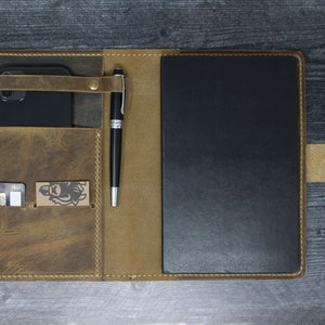 May include: A brown leather notebook cover with a pen holder, a phone pocket, and a card slot. The cover is open to reveal a black notebook.