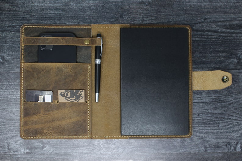 May include: A brown leather notebook cover with a pen holder, a phone pocket, and a card slot. The cover is open to reveal a black notebook.