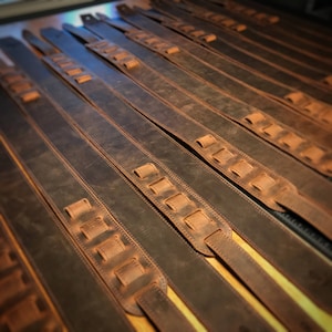 May include: Multiple brown leather guitar straps with adjustable buckles and stitching. The straps are laid out in rows on a surface.
