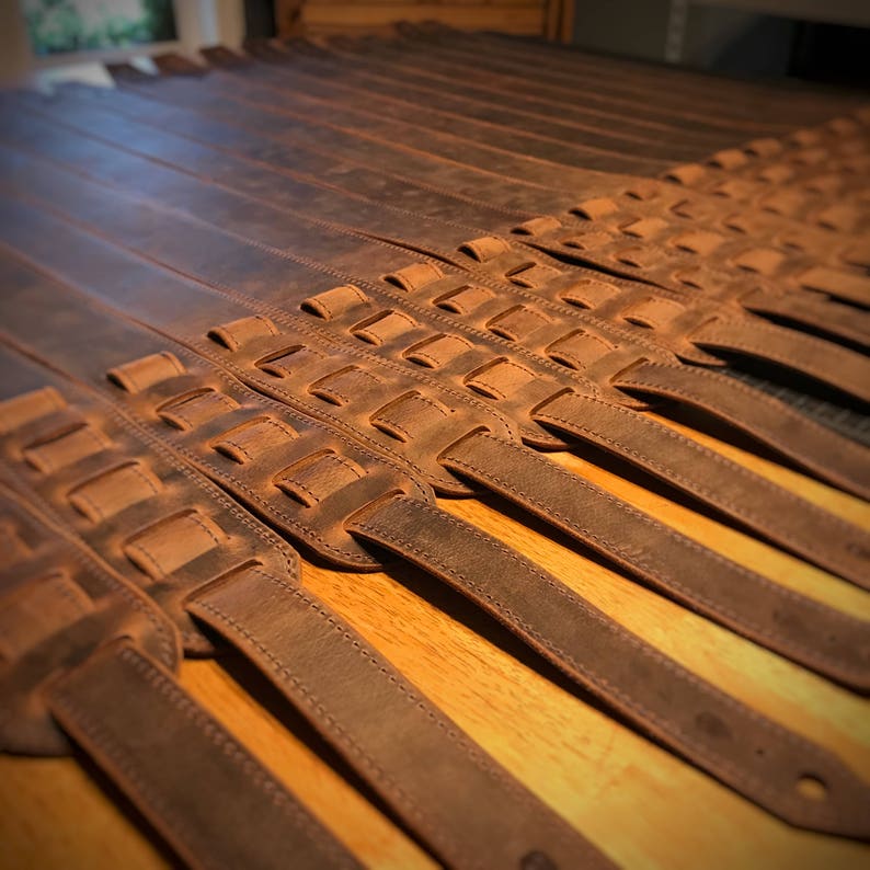 May include: A group of brown leather guitar straps with a woven design. The straps are laid out in rows on a wooden surface.