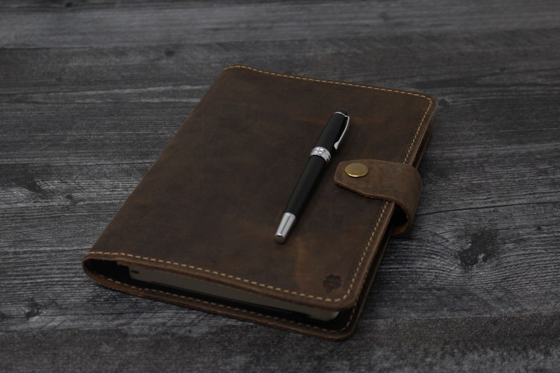 May include: A brown leather-bound journal with a pen on top. The journal has a flap closure with a brass button.