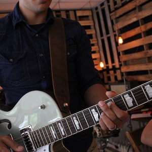 May include: A person playing a light blue electric guitar with a brown leather strap. The guitar has a white pickguard and a black fretboard.