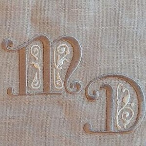 Könnte beinhalten: Zwei gestickte Buchstaben, "M" und "D", in einer traditionellen Schriftart mit dekorativen Verzierungen. Die Buchstaben sind mit braunem Garn auf einem beigefarbenen Leinenstoff gestickt.