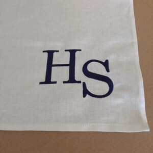 Könnte beinhalten: Cremefarbenes Leinenhandtuch mit dunkelblauen, gestickten Initialen "HS". Das Handtuch ist gefaltet und zeigt eine Ecke mit den gestickten Buchstaben. Der Stoff hat eine feine Textur.