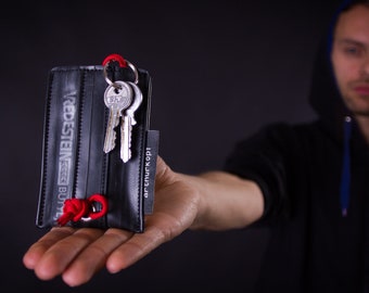 May include: A black leather key holder made from recycled bicycle tire inner tubes. The key holder has a red string loop and the text "Vredestein BT" printed on the front. The key holder is being held in a person's hand.