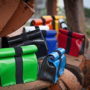 Può includere: Una fila di sacche per il pranzo colorate realizzate con camere d'aria riciclate. Le borse sono di vari colori, tra cui verde, nero, blu, rosso e arancione. Le borse hanno un design semplice con una patta che si ripiega e un cinturino nero.