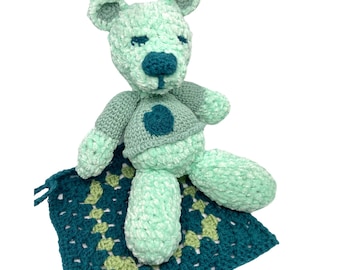 May include: A mint green crocheted teddy bear with a teal nose and a teal heart-shaped patch on its chest. The bear is sitting on a teal and green crocheted blanket. The bear's arms and legs are outstretched.