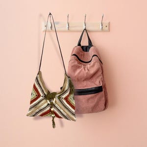 May include: A crocheted shoulder bag with brown, green, and white stripes and a tassel hanging from the bottom. The bag is hanging on a hook next to a pink corduroy backpack.