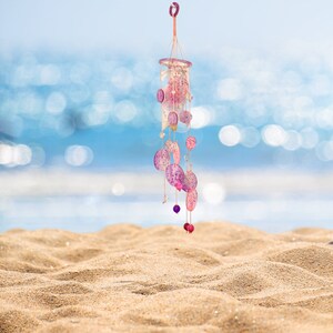 May include: A decorative wind chime with a circular top and dangling, oval-shaped ornaments in shades of pink and purple. The chime hangs from a string and features small beads. The background shows a blurred ocean and sand.