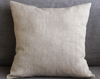 May include: A plain, light brown linen pillow with a subtle texture. The pillow is sitting on a gray couch.