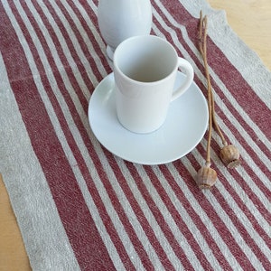 May include: A white coffee cup and saucer on a red and white striped linen table runner.  Dried poppy seed pods are on the table runner.