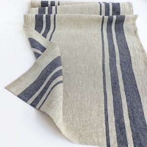 May include: A natural linen table runner with wide blue stripes. The runner is folded and has a textured, woven appearance.