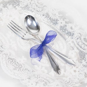 May include: A silver fork and spoon tied together with a blue ribbon, resting on a white lace doily. The silverware is clean and shiny, reflecting the light. The doily has a delicate, intricate pattern.