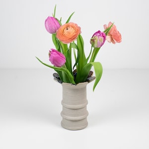 Puede incluir: Un jarrón de cerámica con un acabado texturizado gris claro contiene un vibrante ramo de flores. El arreglo incluye tulipanes rosas y ranúnculos naranjas con hojas verdes, creando una exhibición colorida. El jarrón tiene un diseño único y acanalado.