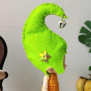 Könnte beinhalten: Eine grüne Filz-Gnommütze mit einer Glocke und Blumenschmuck. Der Gnom trägt ein orangefarbenes Häkelkleid und hat einen hellbraunen Körper.