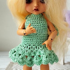 Puede incluir: Una muñeca con pelo largo y rubio que lleva un vestido verde de ganchillo. La muñeca está de pie sobre una alfombra estampada frente a una silla morada. El vestido tiene un corpiño ajustado y una falda acampanada. La piel de la muñeca es clara.