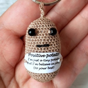 May include: A crocheted keychain featuring a tan potato-shaped figure with black eyes and a stitched mouth. A small white tag displays the text "Positive potato" and an inspirational message. The keychain includes a silver-colored ring.