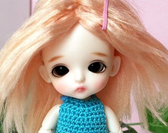 May include: A small doll with pale skin and large dark eyes, featuring shoulder-length peach-colored hair held back by a small pink clip. The doll is wearing a sleeveless, textured turquoise blue crocheted dress that reaches mid-thigh.