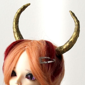 May include: A doll with short orange hair featuring red highlights and a silver hair clip. The doll wears two textured gold-colored horns on its head, purple eyes, and a silver chain necklace with black and silver beads over a black top.