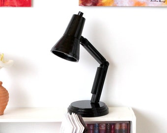 May include: A white desk with a black desk lamp, a book, a vase with white flowers, and a small bookshelf with books. The desk is in front of a white wall with two paintings.
