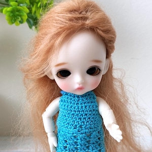 May include: A small doll with long, light brown hair and dark eyes wearing a turquoise crocheted dress. The doll is standing on a patterned surface, with a small wooden bench and greenery in the background.