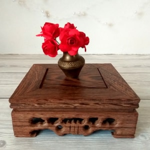 May include: A small, square wooden stand with intricate carvings on the bottom. A brass vase with red silk flowers sits on the stand.