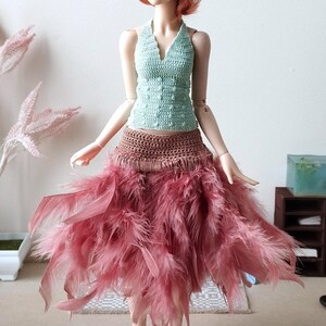 May include: A doll wearing a light green crocheted top and a pink feather skirt. The doll has long, red hair and blue eyes. The doll is standing on a white surface in front of a white wall.