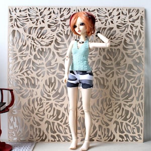May include: A doll wearing a light blue top and camouflage shorts stands in front of a wooden panel with a leaf pattern. The doll has red hair and is accessorized with a necklace. A red chair and a small table are also in the scene.