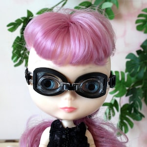 May include: A pink haired doll wearing black goggles with clear lenses. The doll is wearing a black dress with a beaded neckline.